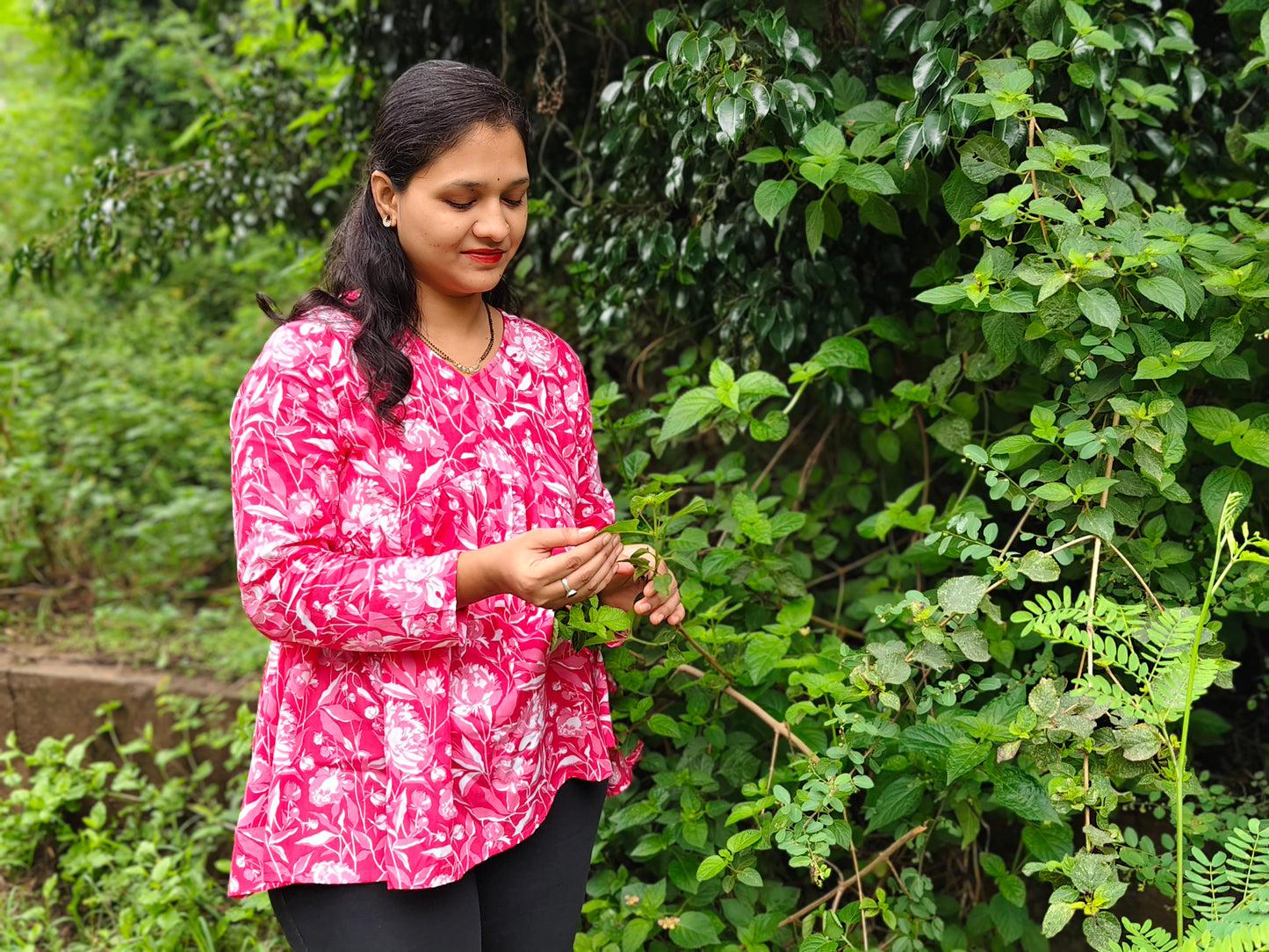 Woman wearing a pink peplum top with floral patterns while standing in a lush green garden